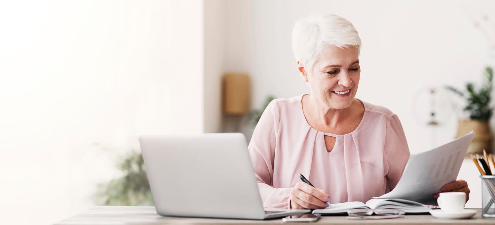 Mulher sorridente lendo e fazendo anotações em um caderno aberto, com um laptop e papéis sobre a mesa