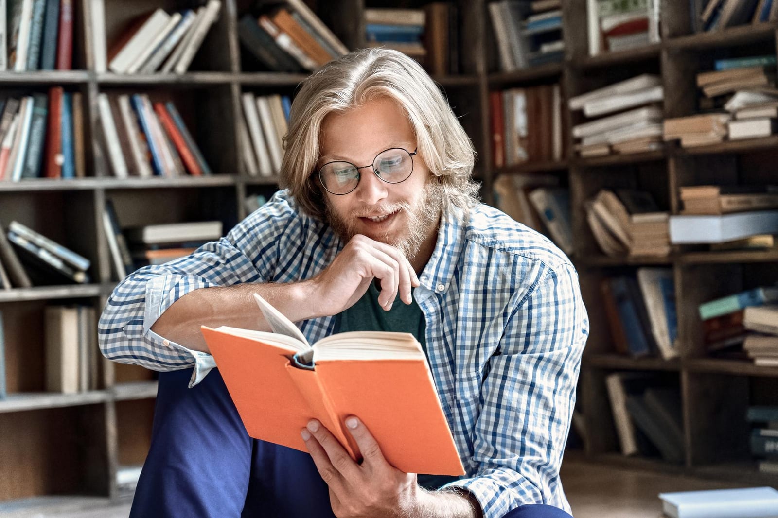 Homem concentrado lendo um livro de capa colorida em frente a estantes repletas de livros